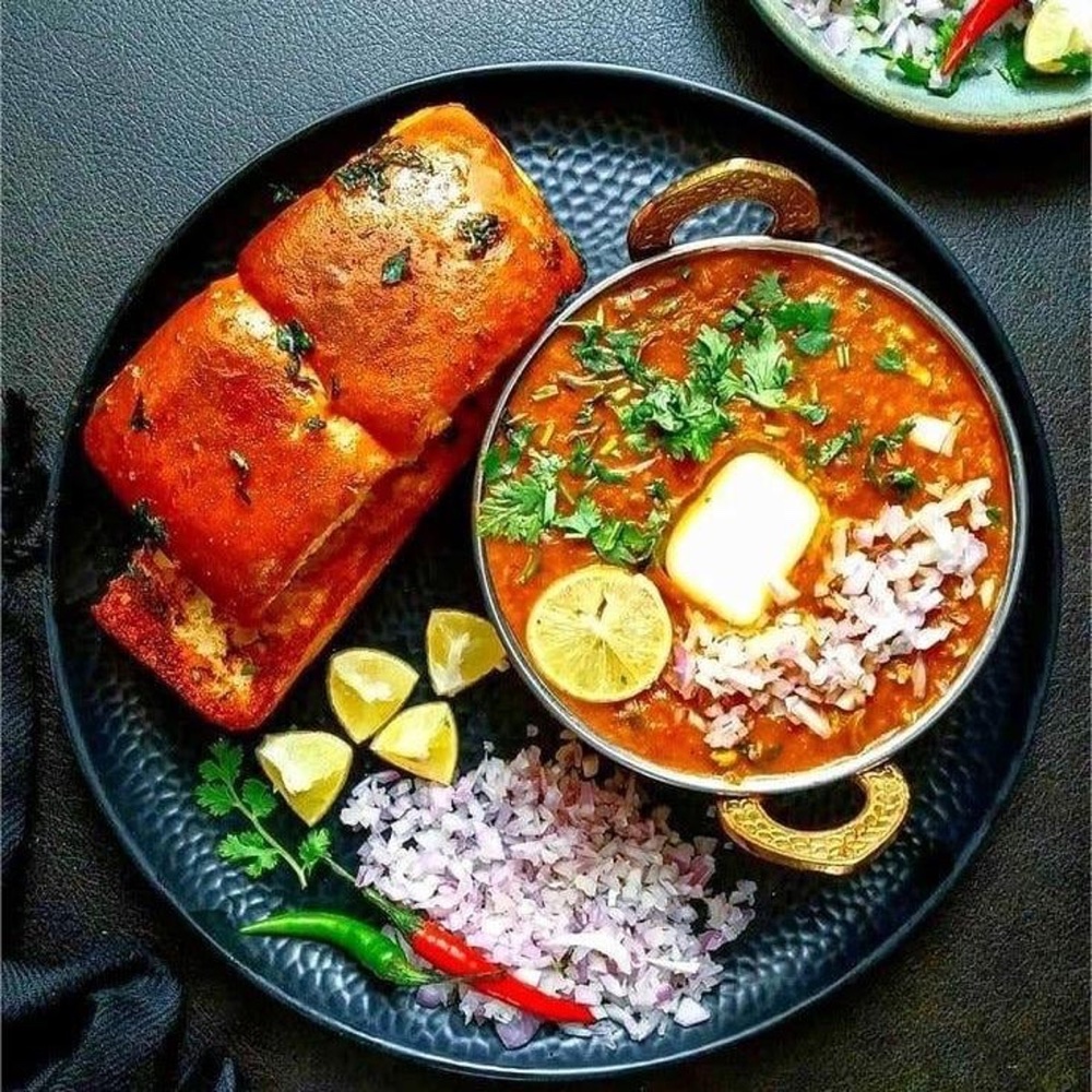Paav bhaji with buttered pav, chopped onion and lime on a black plate